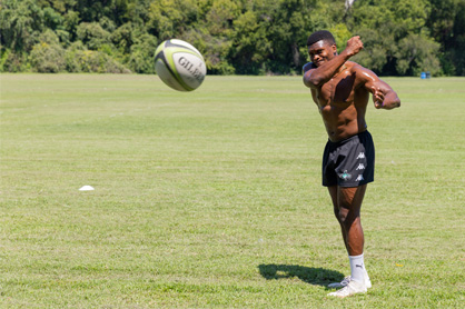 Ani sits in a black polo talking to the camera in front of a stadium backdrop.
