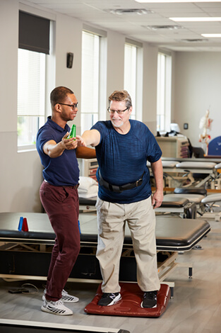 Patient and therapist are standing together while patient is performing exercise with one arm extended forward