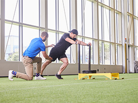 Athlete is pushing a sled across a field with the supervision of a trainer