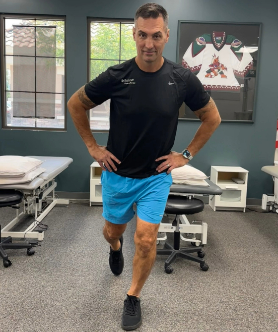 A male physical therapist with his hands on his hips and his left leg out in front of him. 