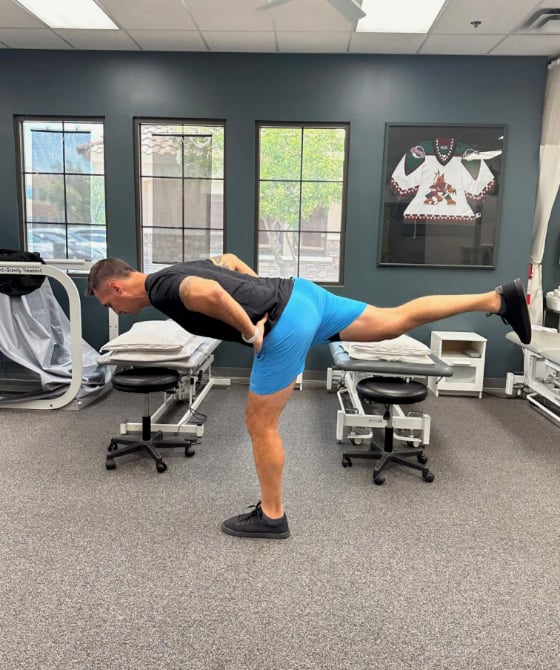 A male physical therapist with his hands on his hips with his left leg forward as he leans forward. 