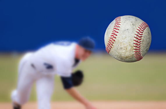 Pitcher is throwing a baseball towards camera. Baseball is close to the camera and pitcher is in the background blurred out