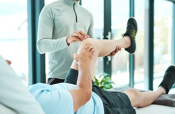 Patient laying on back with one leg in the air while therapist  checks their knee
