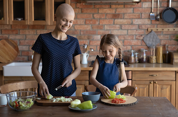 Cancer survivor is in the kitchen cutting vegetables with her daughter