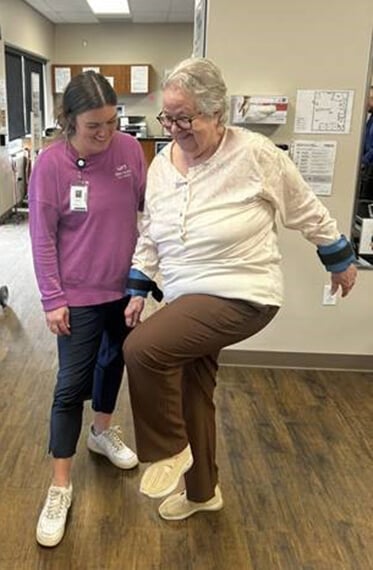 Karen balancing on one leg with the support of physical therapist Kelsey