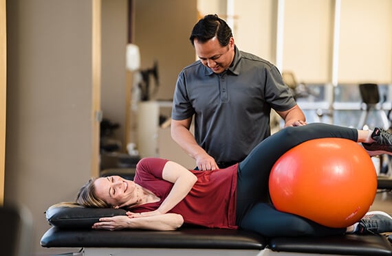 Patient laying on her side on a table with an exercise ball in between her feet. A therapist is beside her
