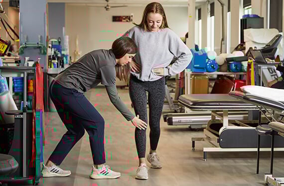 Patient is taking one step towards the camera as a physical therapist is bending over and touching their knee