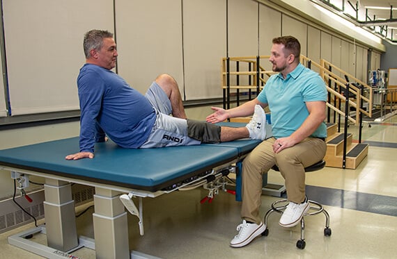 Patient is sitting on an examination table with an ice pack on one knee while talking with a physical therapist that is sitting beside the table