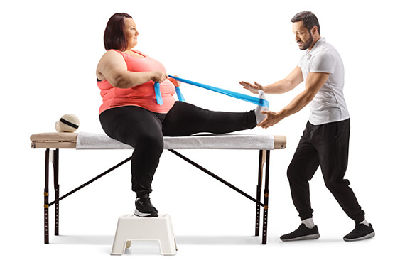 A woman is sitting on a table while using a band to stretch her hamstrings. Her therapist is beside her lifting her foot up